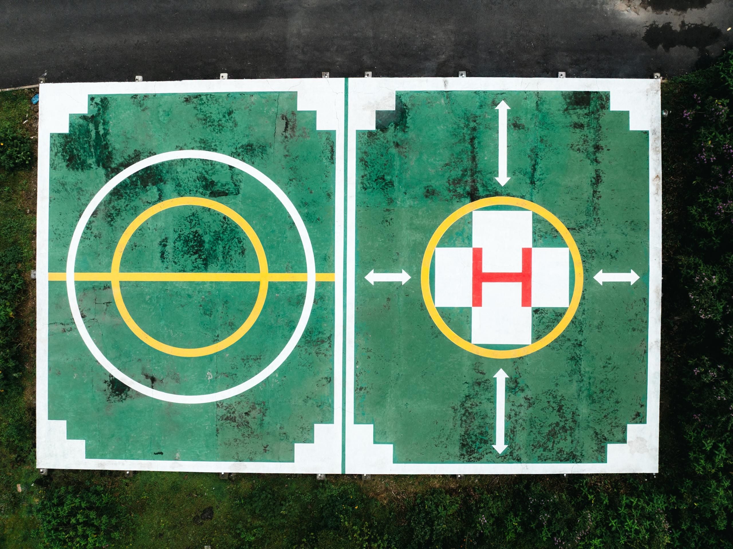 A basketball court with a hoop and a H symbol, surrounded by a green surface with white and yellow markings.