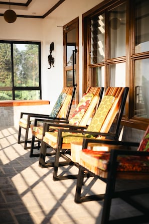 A room with a row of wooden chairs with colorful cushions, large windows with wooden frames, and a hanging light fixture.