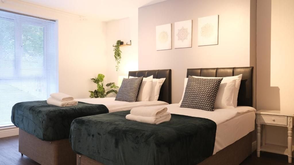 A hotel room with two beds, a bedside table, a window with blinds, and wall art.