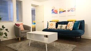 Een moderne woonkamer met een grijze fauteuil, een witte salontafel en een blauwe bank met gekleurde kussens.