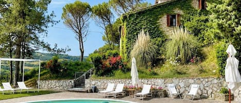 A swimming pool with lounge chairs and umbrellas, surrounded by greenery and a stone wall.