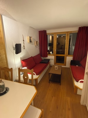 Ein gemütliches Wohnzimmer mit einem roten Sofa, Holzstühlen, einem kleinen Tisch und einem Fenster mit Vorhängen.