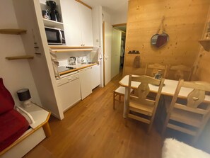 Una cocina compacta con armarios blancos, un microondas y un lavamanos. Hay un comedor con una mesa y sillas de madera.