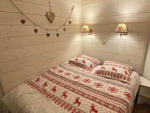 Ein Zimmer mit einer holzverkleideten Wand, einem Bett mit gemusterten Betttextilien, zwei Wandlampen und herzförmigen Dekorationen.