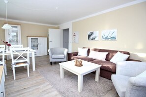 A modern living room with a white coffee table, a brown sofa, and a grey armchair. There is a dining table with chairs and a white lamp. - Holiday apartment in Sylt with 3 bedrooms (Sylt \/ OT Westerland)