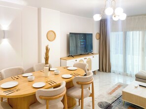 A modern living room with a dining area, a flat-screen TV mounted on a wooden cabinet, and a large window with curtains. - 3BRs Bohimian Citycenter Apartmemt (Cairo Governorate)