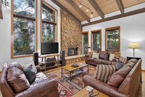 A spacious living room with a stone wall, large windows, a fireplace, leather sofas, and a flat-screen TV. - Spring Hill Hideaway - Vail (Vail)