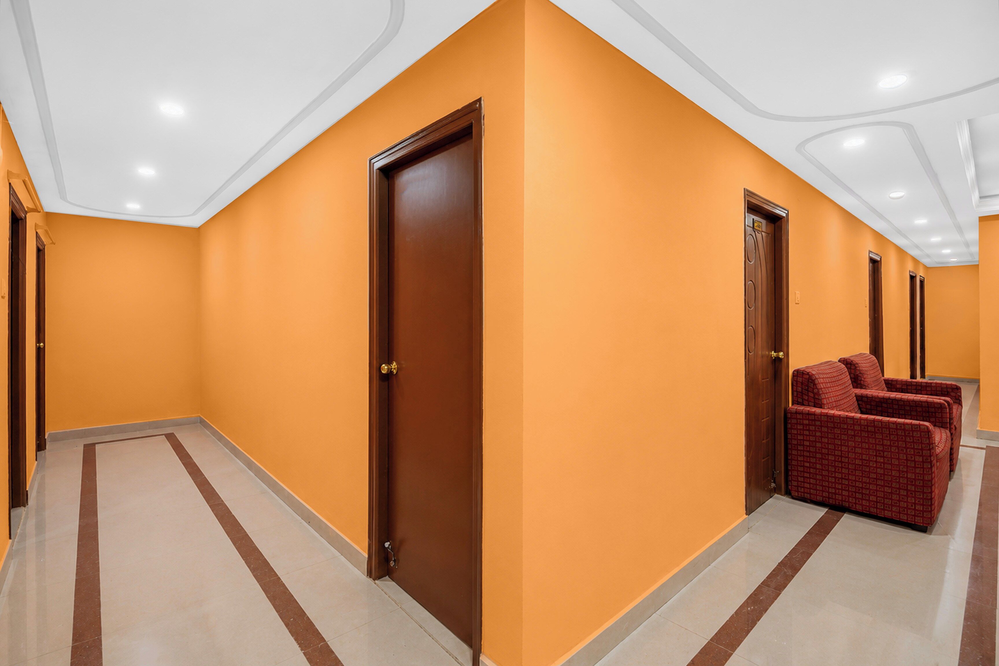 A hotel corridor with orange walls, wooden doors, and a seating area with red upholstered chairs.