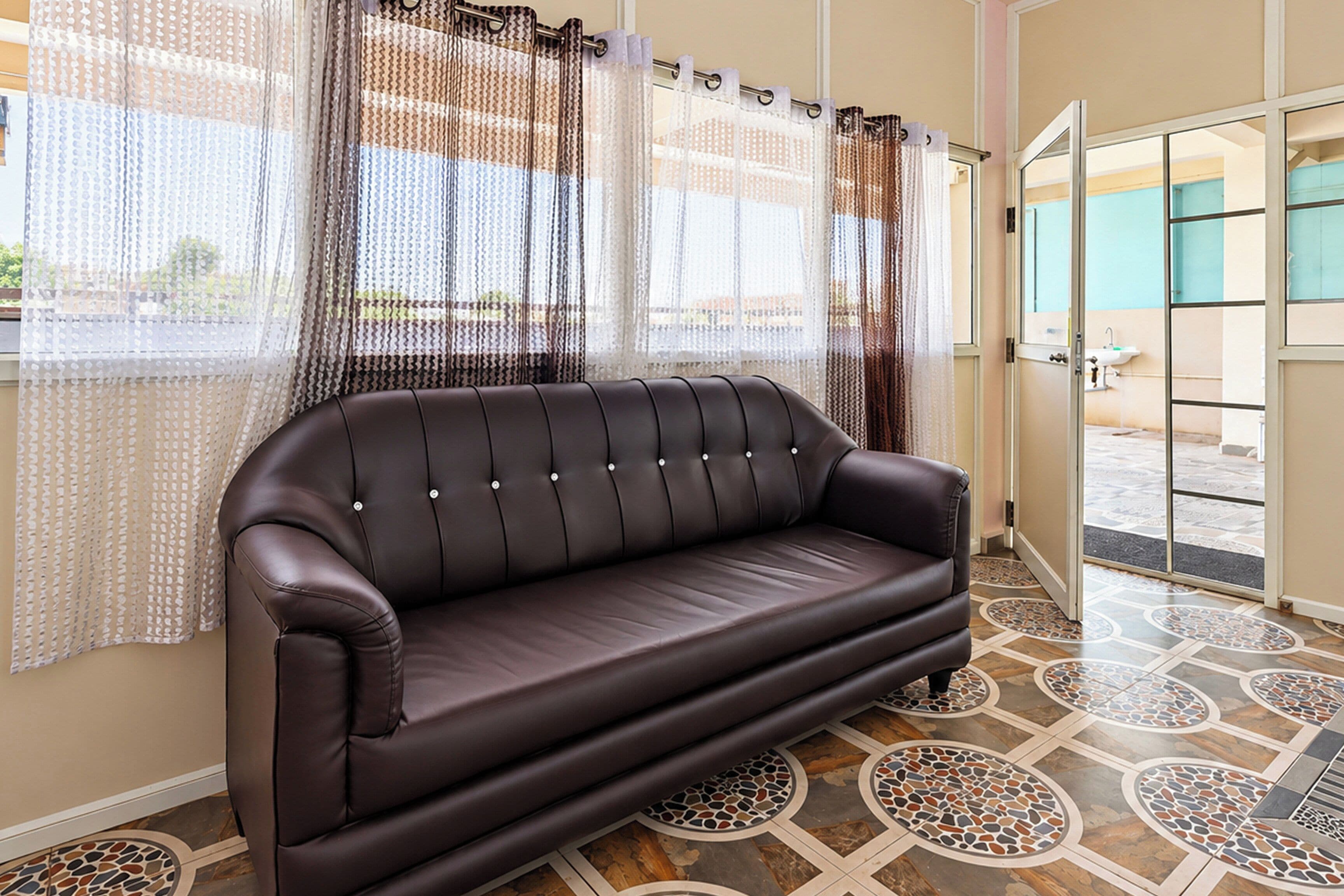 A dark brown leather sofa in a room with patterned floor tiles and sheer curtains with a geometric pattern.