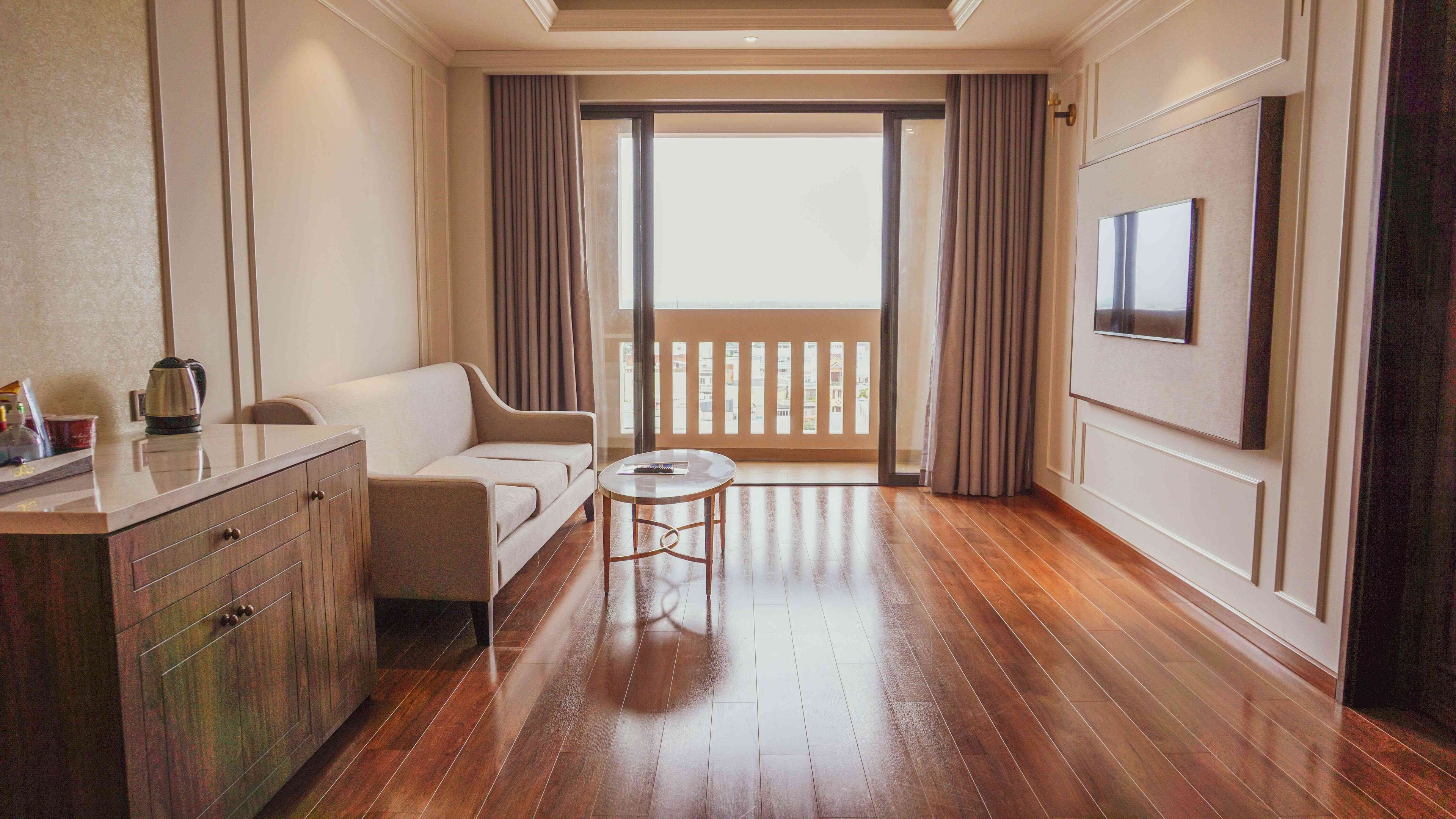 Habitación de hotel con piso de madera, un sofá, una pequeña mesa redonda, un televisor montado en la pared y un ventanal corredero que da a un balcón.