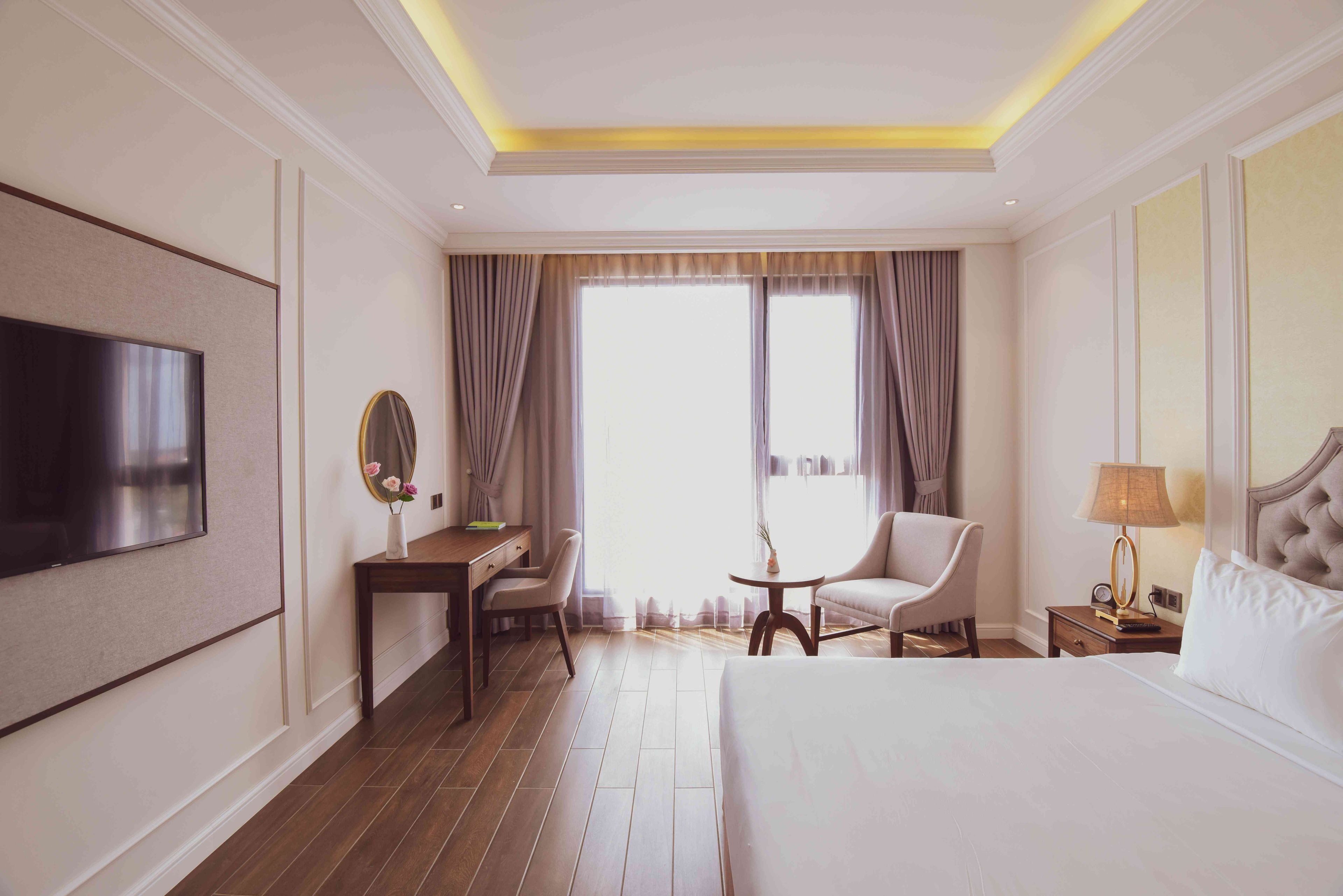 A hotel room with a bed, a desk, a chair, a TV, a bedside table with a lamp, and a window with curtains.