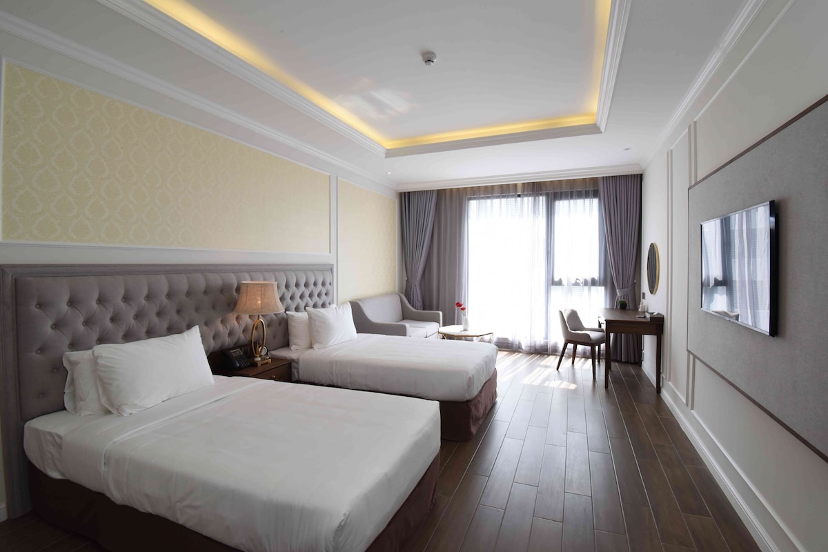 A hotel room with two beds, a desk, a chair, a TV, and a window with curtains.