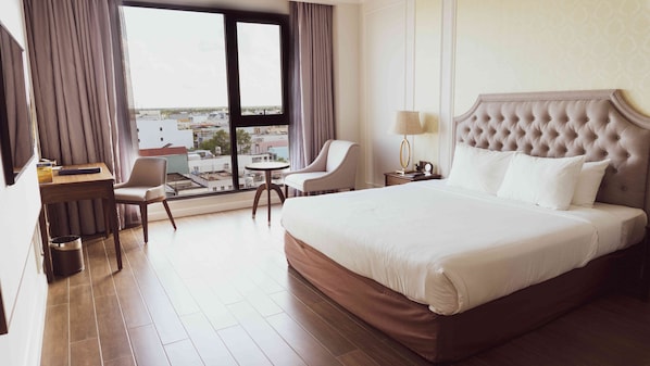 A hotel room with a large bed, a desk with a chair, a small table, and a view of the city through large windows.