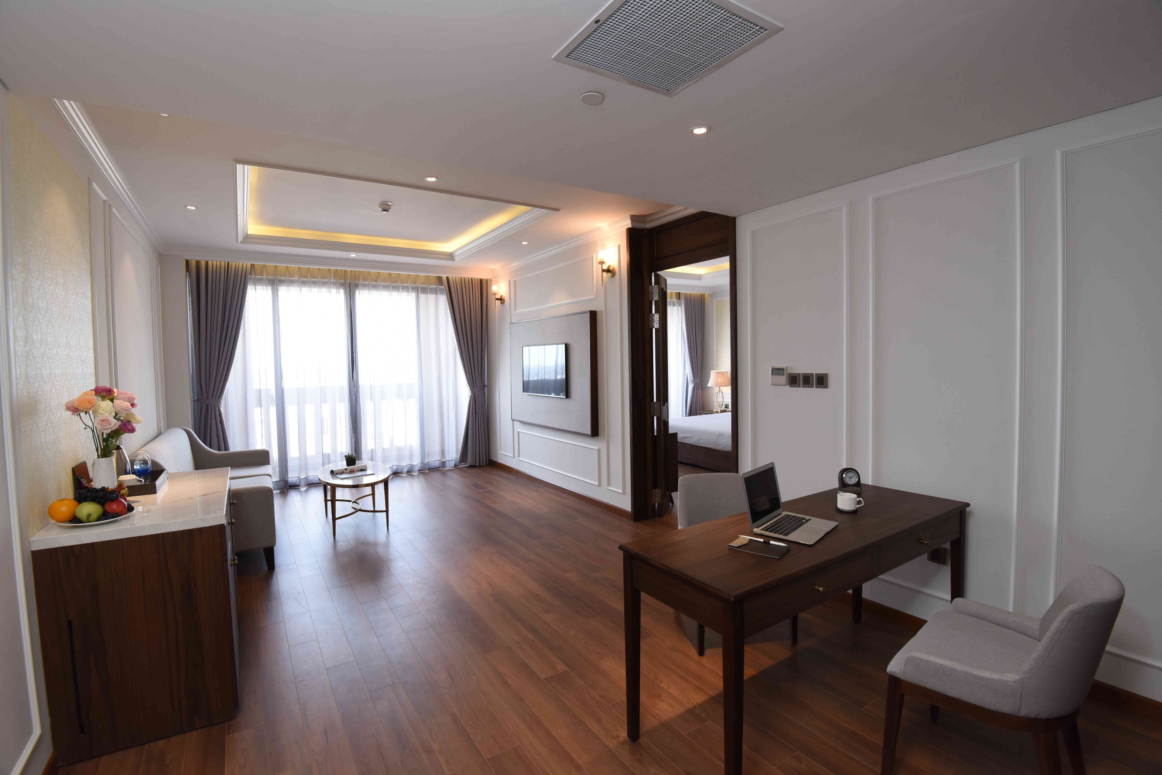 A hotel room with a wooden floor, a desk with a laptop, a chair, a TV mounted on the wall, and a bed visible through an open door.