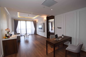 A hotel room with a wooden floor, a desk with a laptop, a chair, a TV mounted on the wall, and a bed visible through an open door. - Phu Cuong Hotel Ca Mau (An Xuyen)