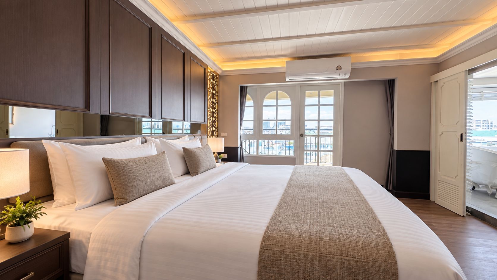 A hotel room with a large bed, a nightstand with a lamp, a potted plant, and a view of the city through the windows.