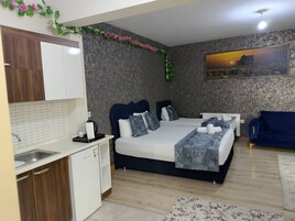 Habitación de hotel compacta con kitchenette, cama con cubrecama gris, sofá azul y un cuadro en la pared.
