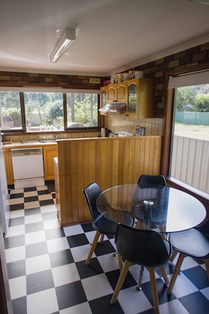 A compact kitchen with a checkered floor, wooden cabinets, a dining table with chairs, and a view of the outdoors through a window. - The Gap - Family Nature Retreat (Halls Gap)