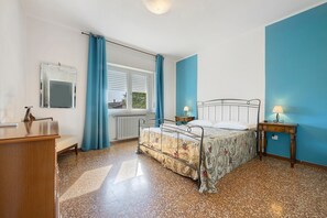 A bedroom with a bed, bedside tables, a desk, a chair, a radiator, and a window with curtains. - Apartment 'La Prua - Capitan Libeccio' with Lake View, Wi-Fi, and Air Conditioning (Garda)