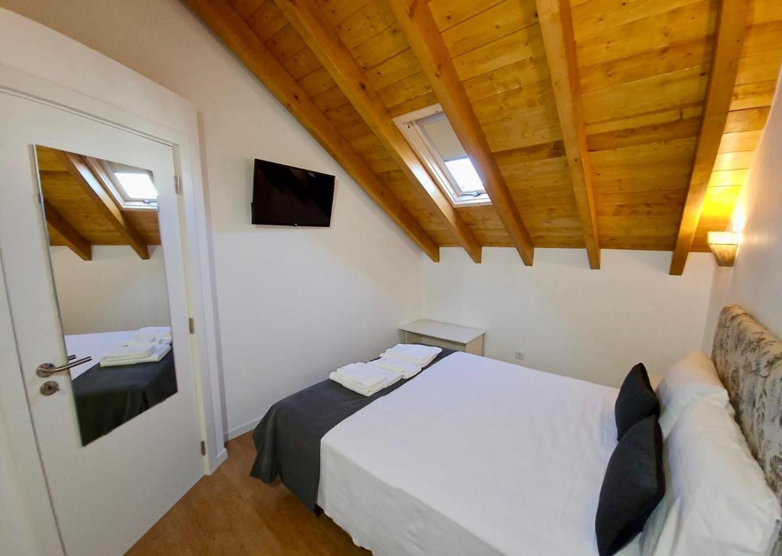 A bedroom with a wooden ceiling, a bed with white bedding, a small bedside table, and a television on the wall.