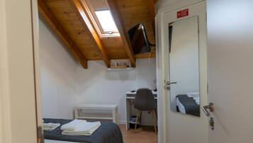 Una camera da letto con un letto, una scrivania, una sedia, una TV montata a soffitto e una porta con un cartello rosso.
