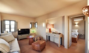 Una sala moderna con televisión de pantalla plana, un sofá, una mesa de centro y una mesita auxiliar. Hay una lámpara sobre la mesita y una cama en la habitación contigua.