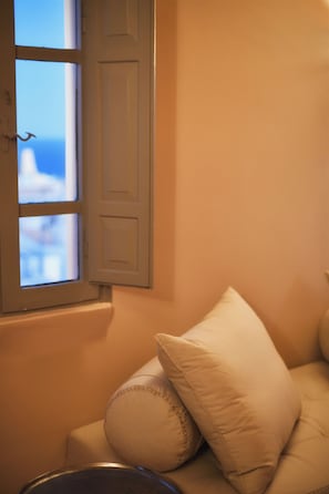 A room with a window, a white door, and a beige sofa with pillows. - Terra Verde Oia Suites - Therra Grand Suite Sea View (Oia)
