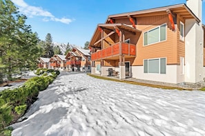 Una hilera de cabañas de madera con balcones, suelo cubierto de nieve y montañas al fondo.