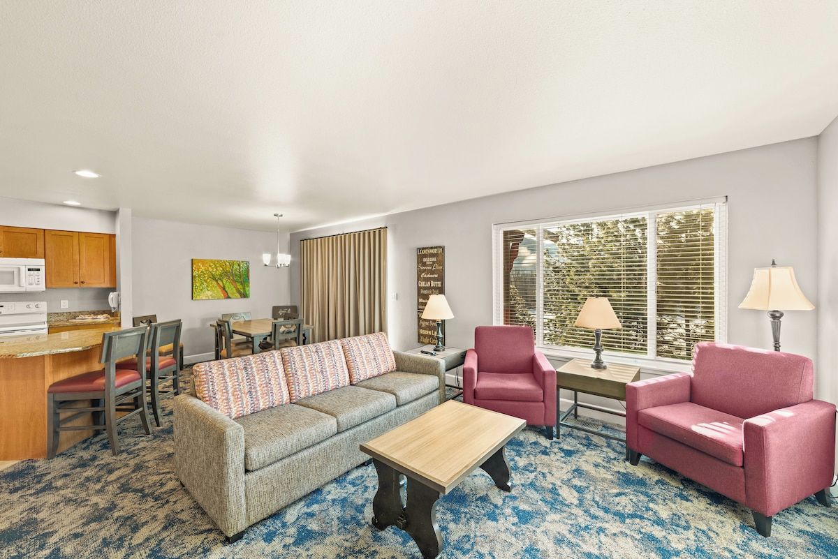 Ein Wohnzimmer mit einer Couch, zwei Sesseln, einem Couchtisch, einem Essbereich mit Stühlen, einer Küchenzeile und einem großen Fenster mit Jalousien.