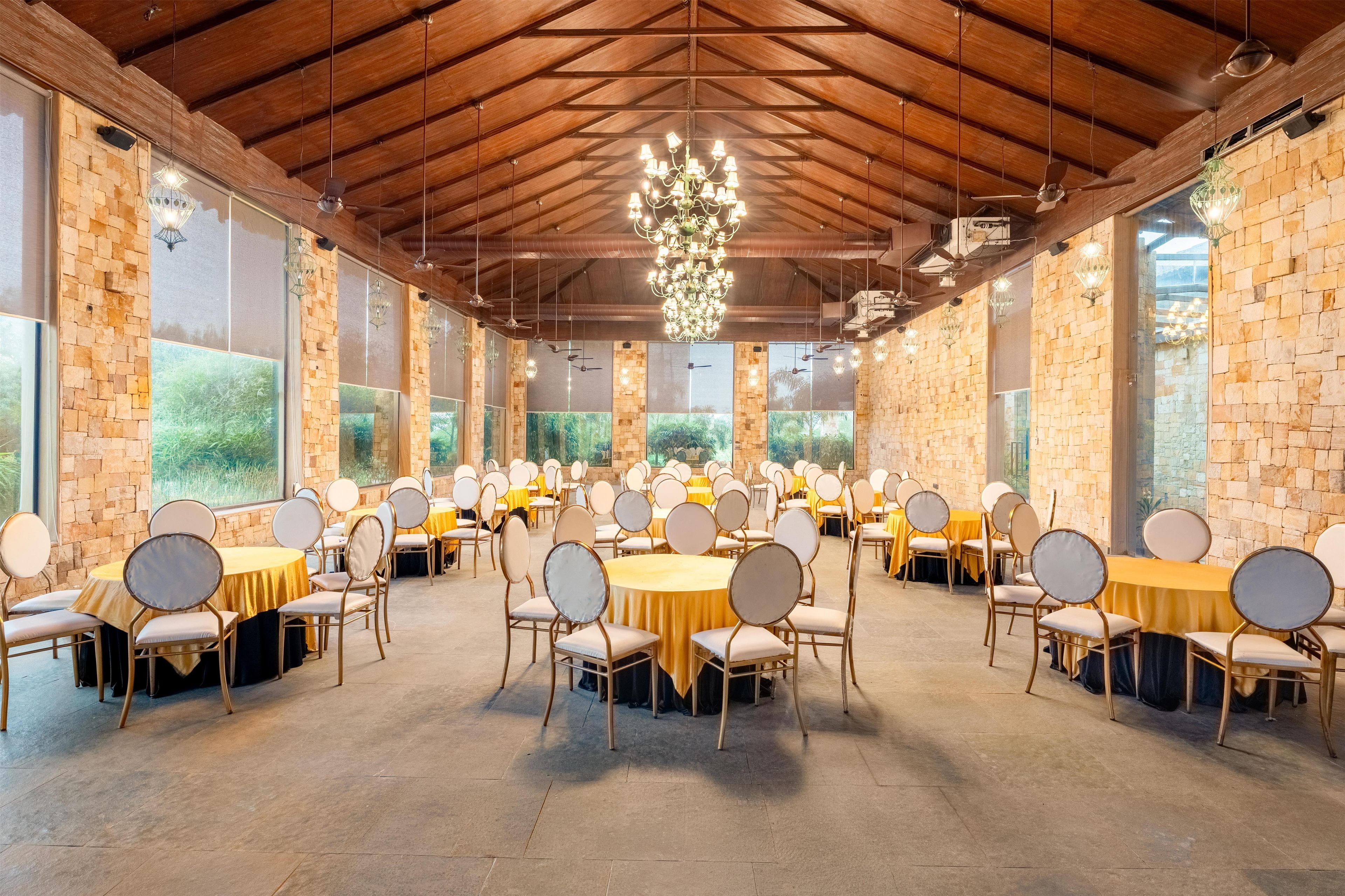 A spacious hall with multiple round tables and chairs, large windows, and a high ceiling with exposed beams.