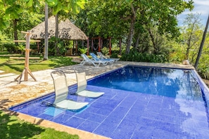 Piscina com azulejos azuis, cercada por espreguiçadeiras e uma estrutura com telhado de palha.