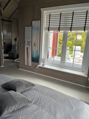 Een slaapkamer met een groot bed, een raam met zonwering, een schilderij van een zeilboot en uitzicht op tuinmeubilair.