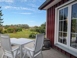 Un patio con mesa y sillas, un edificio rojo con ventanas amplias y un jardín con diversas plantas y flores.