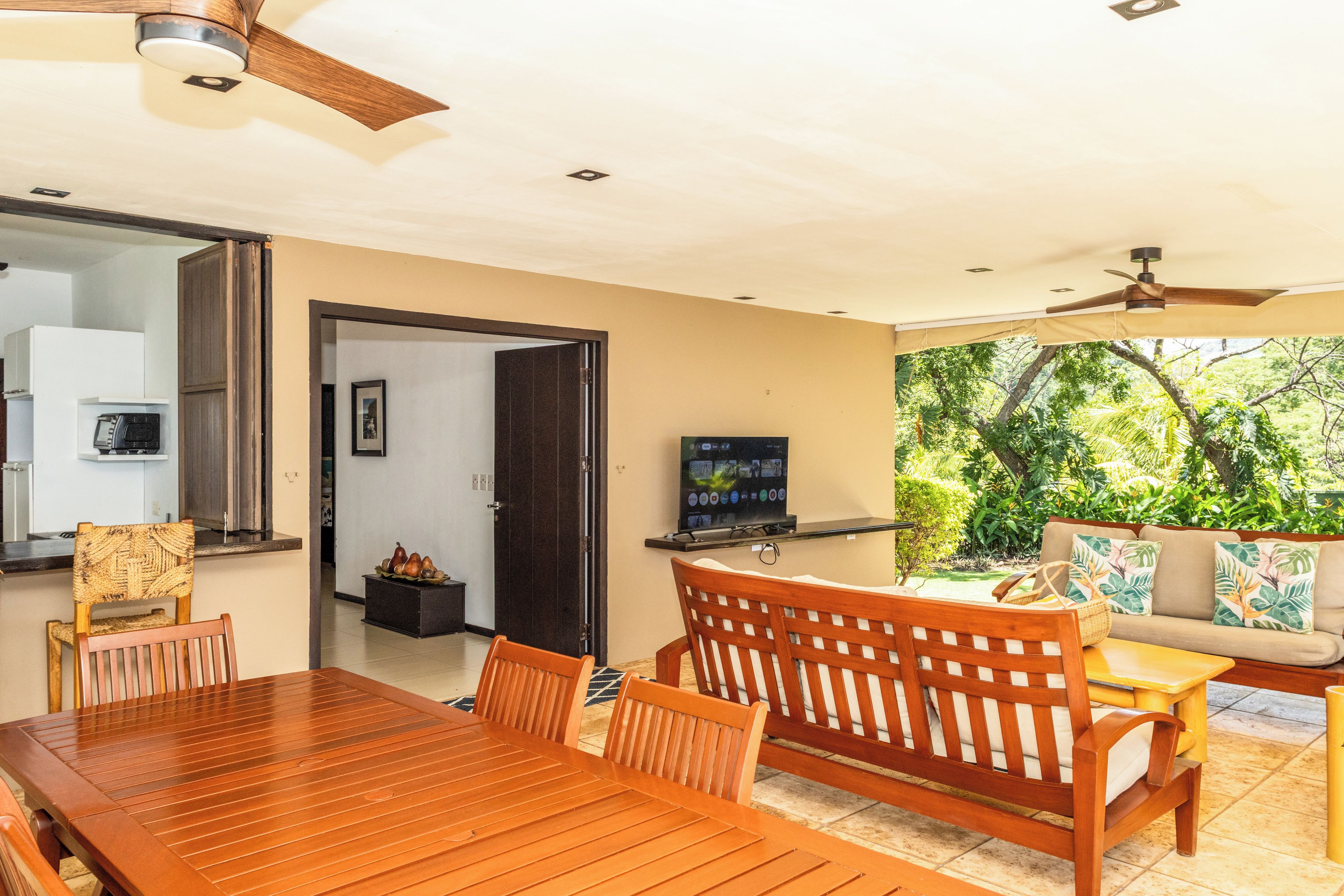 Un comedor con una mesa y sillas de madera, un televisor sobre un mueble, y vista a un jardín por una puerta abierta.