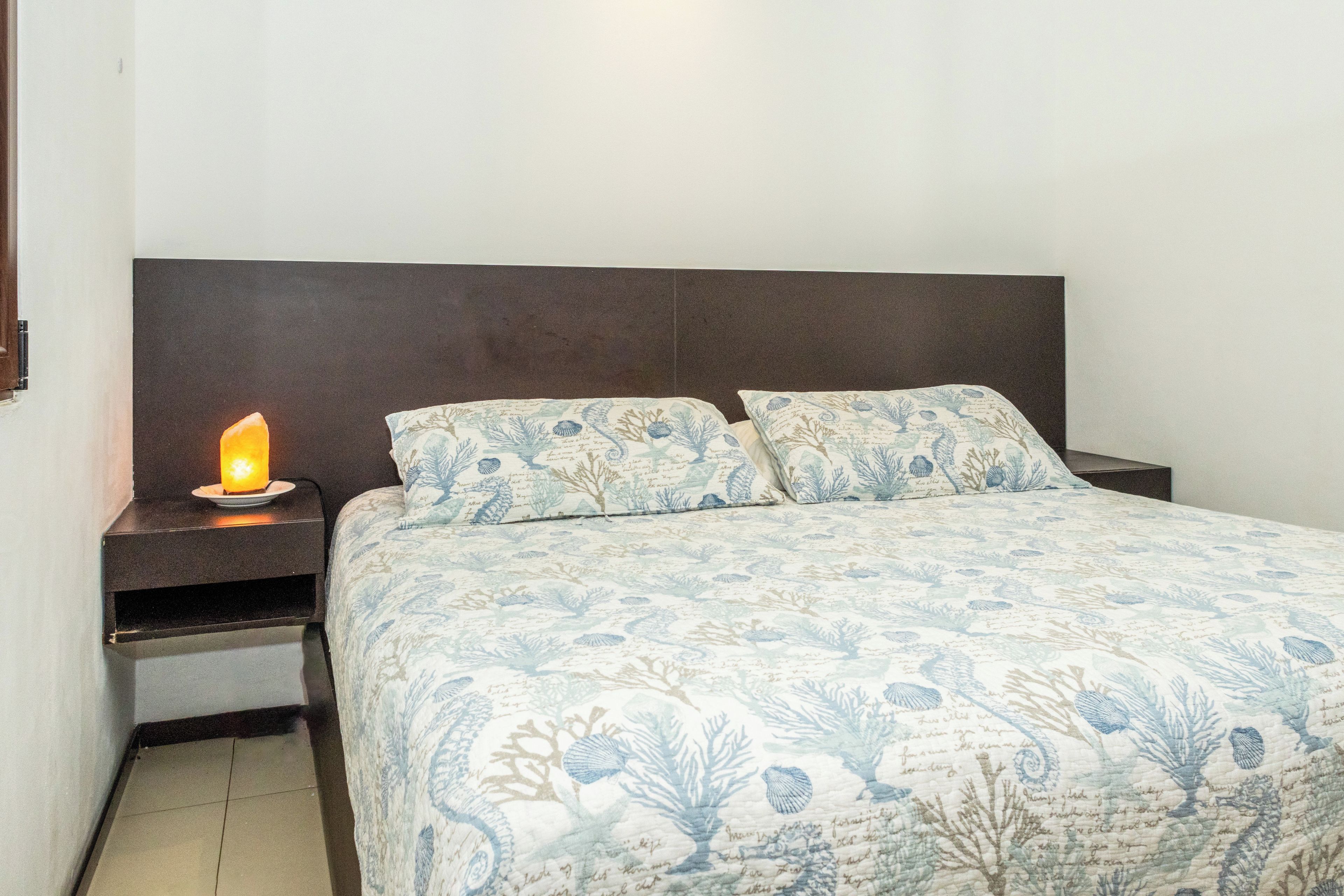 Una cama con colcha estampada con motivos florales, dos almohadas, una mesita de noche con lámpara y un cabecero oscuro.