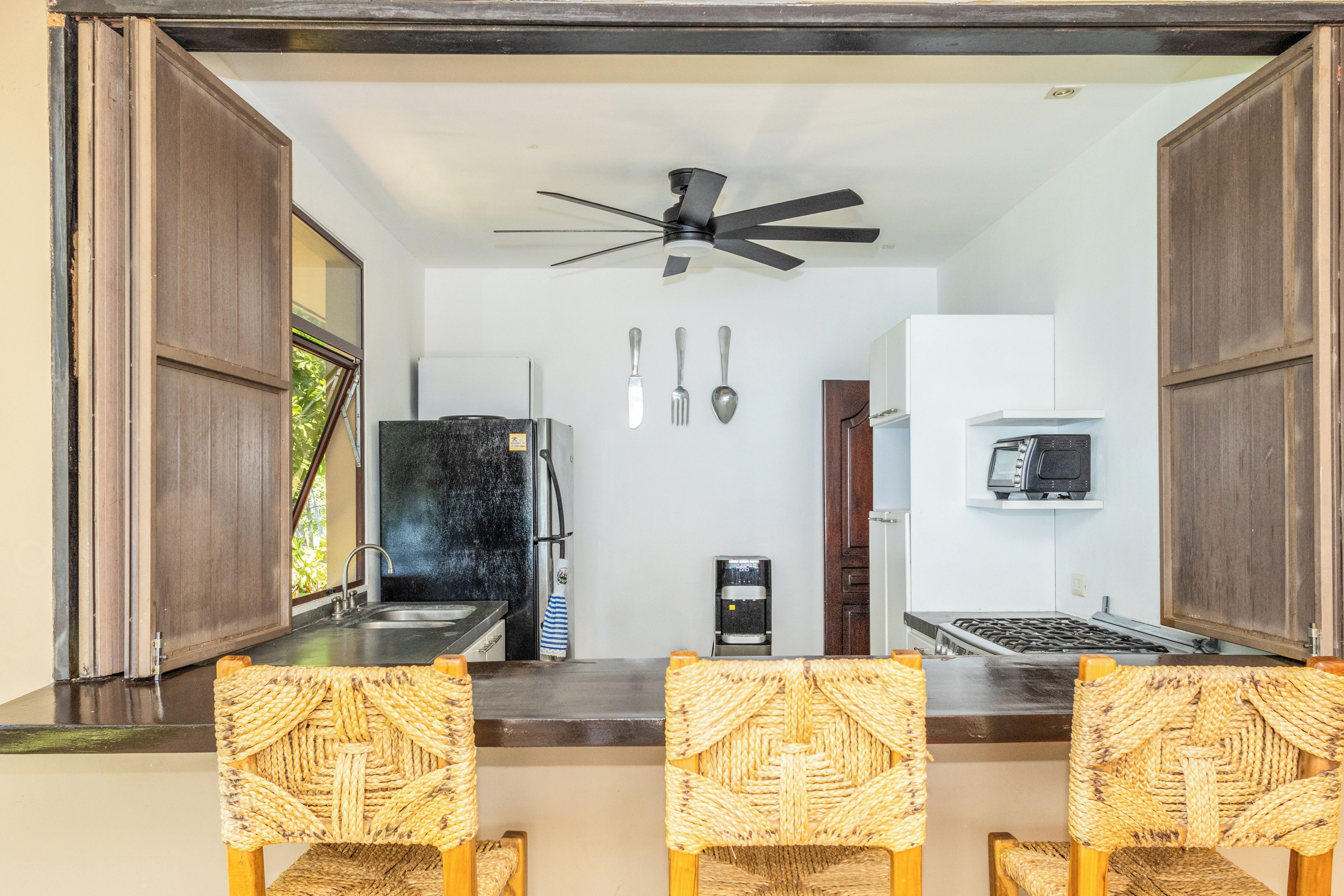 A kitchen with a refrigerator, microwave, and coffee maker. There are three woven chairs and a ceiling fan.