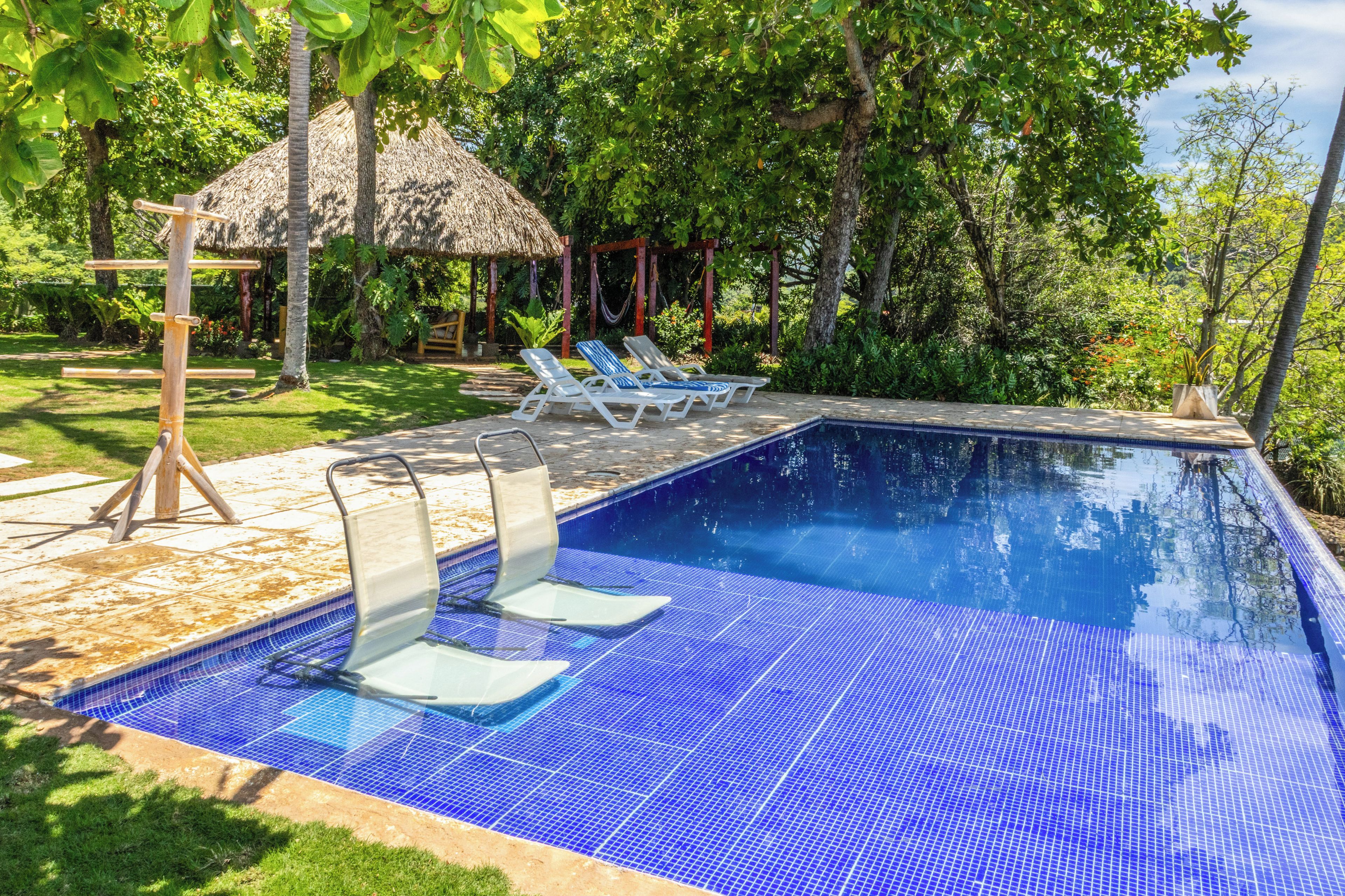 Una piscina con baldosas azules, dos sillas de descanso, una cubierta de madera y una estructura de techo de paja.