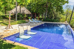 Una piscina con baldosas azules, dos sillas de descanso, una cubierta de madera y una estructura de techo de paja.