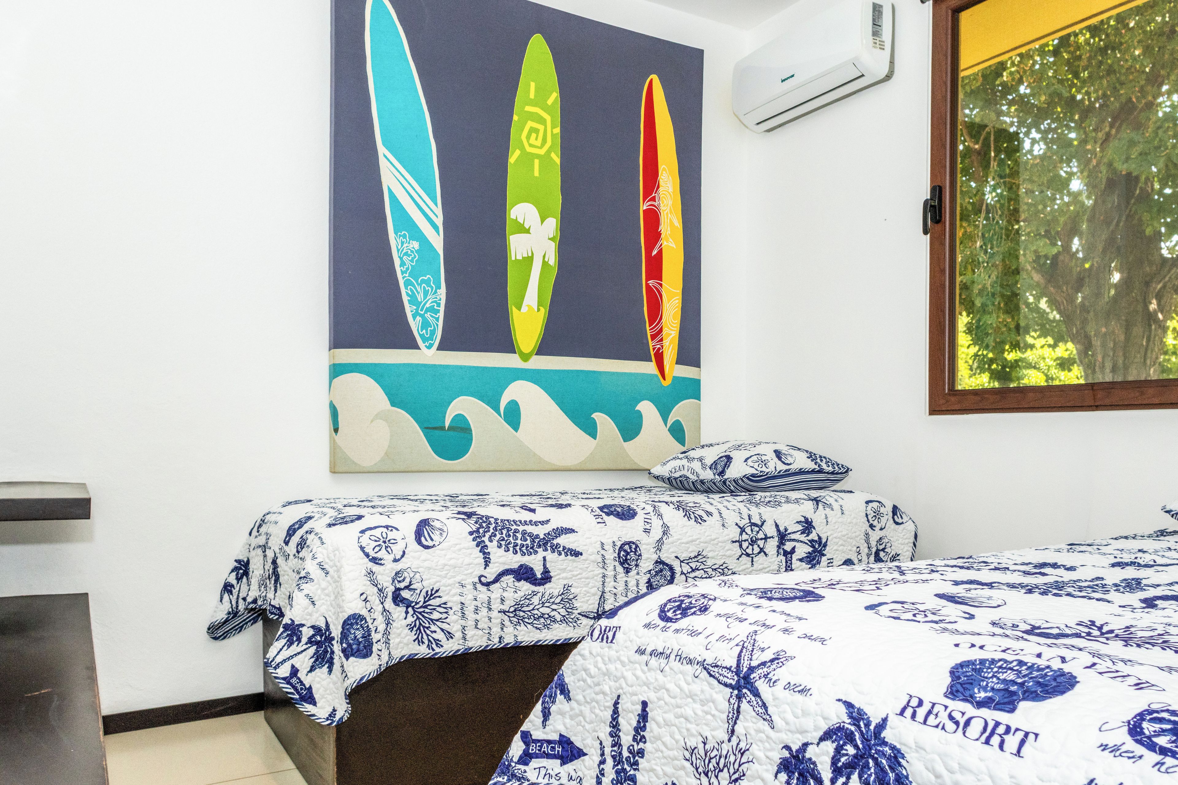 Un dormitorio con dos camas, aire acondicionado de pared y ventana con vista a árboles.