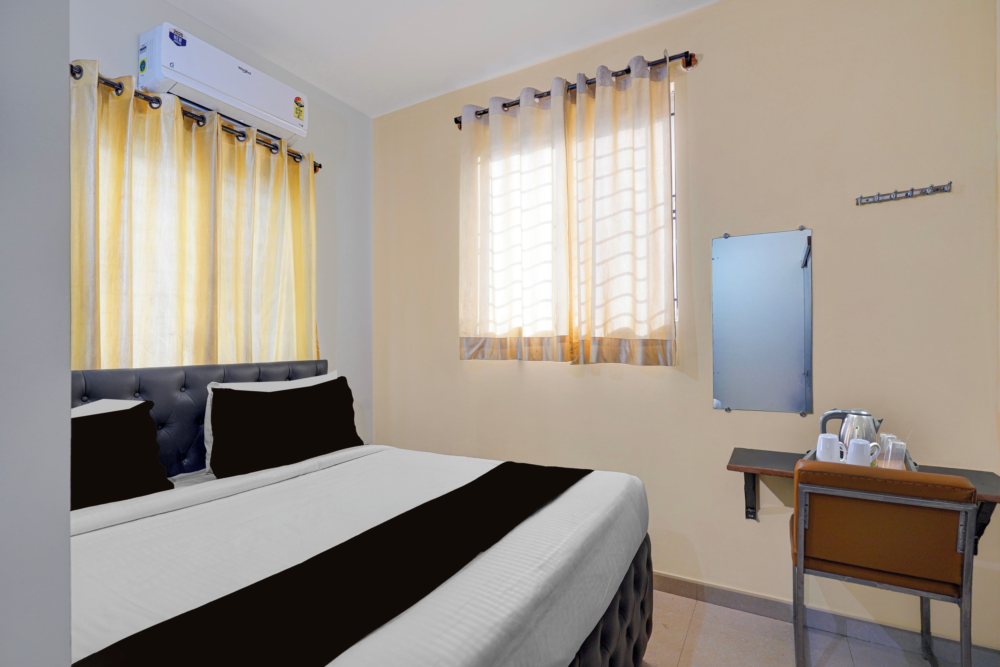 A hotel room with a bed, a desk with a chair, a mirror, and a window with curtains.