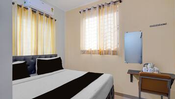 A hotel room with a bed, a desk with a chair, a mirror, and a window with curtains.