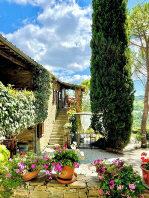 A rustic stone building with a staircase leading to a wooden deck, surrounded by lush greenery and flowering plants. - Apartment with terrace over Umbrian countryside (Gubbio)