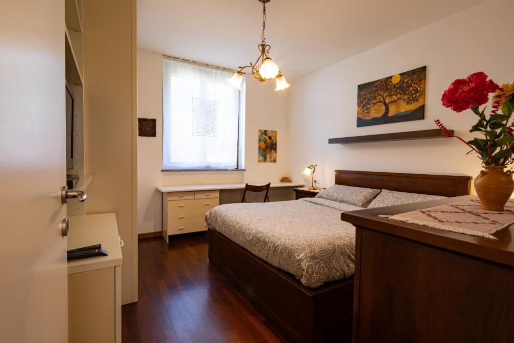 Ein Schlafzimmer mit Bett, Nachttisch, Lampe, Fenster, Kunstwerk und einer Blumenvase.