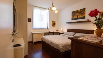 Ein Schlafzimmer mit Bett, Nachttisch, Lampe, Fenster, Kunstwerk und einer Blumenvase.
