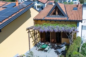 A residential building with a sloped roof, solar panels, a covered patio with outdoor furniture, and a garden with purple flowering plants.