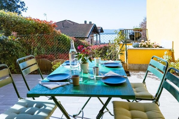 An outdoor dining area with a table set for two, surrounded by chairs, and a view of a building and the sea in the background.
