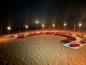 Zona relax al costat de la platja amb fileres de gandules i ombrelles, il·luminada per llums penjants.