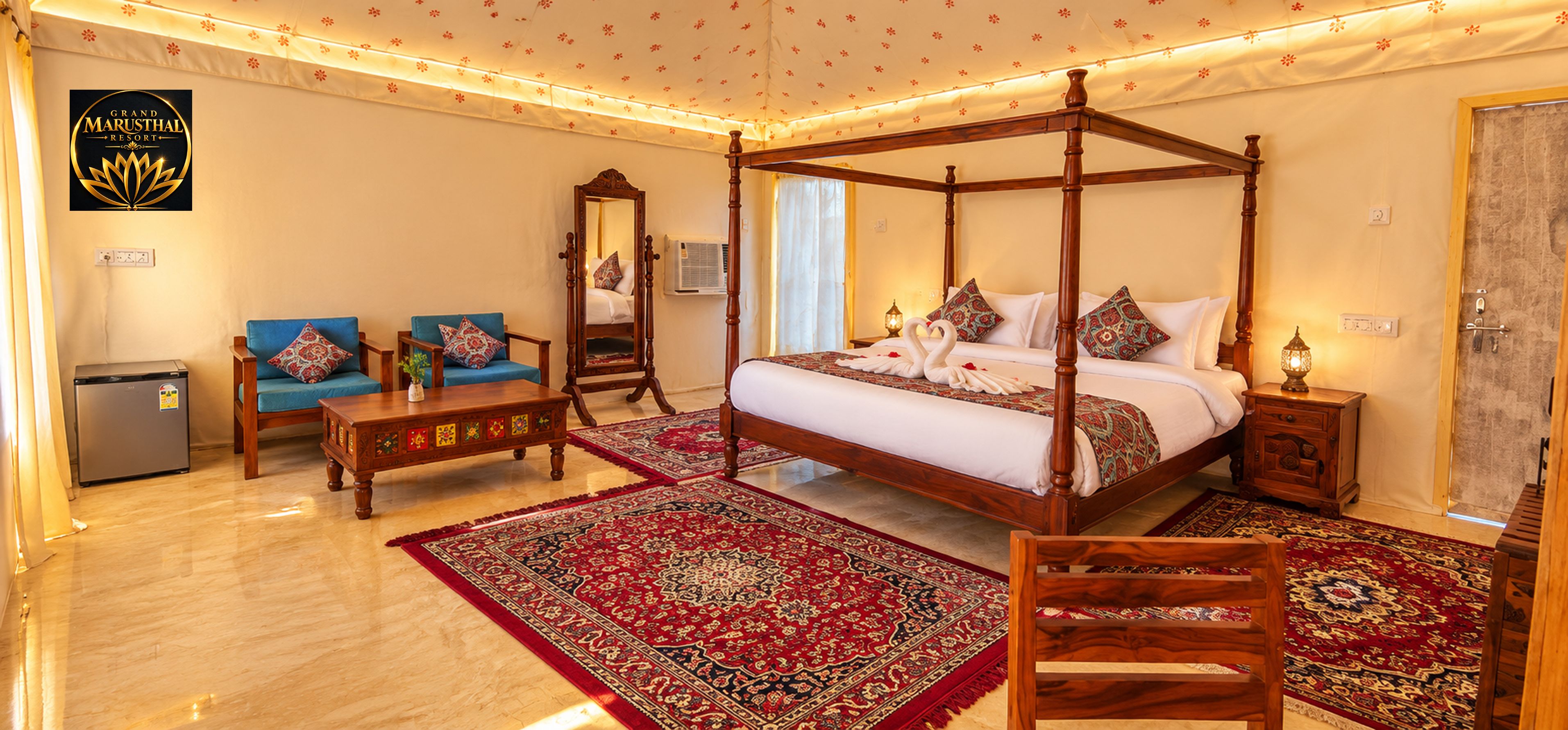 A four-poster bed with a red and black patterned bedspread, a wooden nightstand with a lamp, a blue sofa set with patterned cushions, and a small refrigerator.
