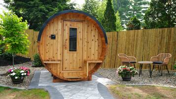 Une structure de sauna en bois, dotée d’une porte, d’un espace détente avec des chaises et une table, ainsi que de plantes en pot.