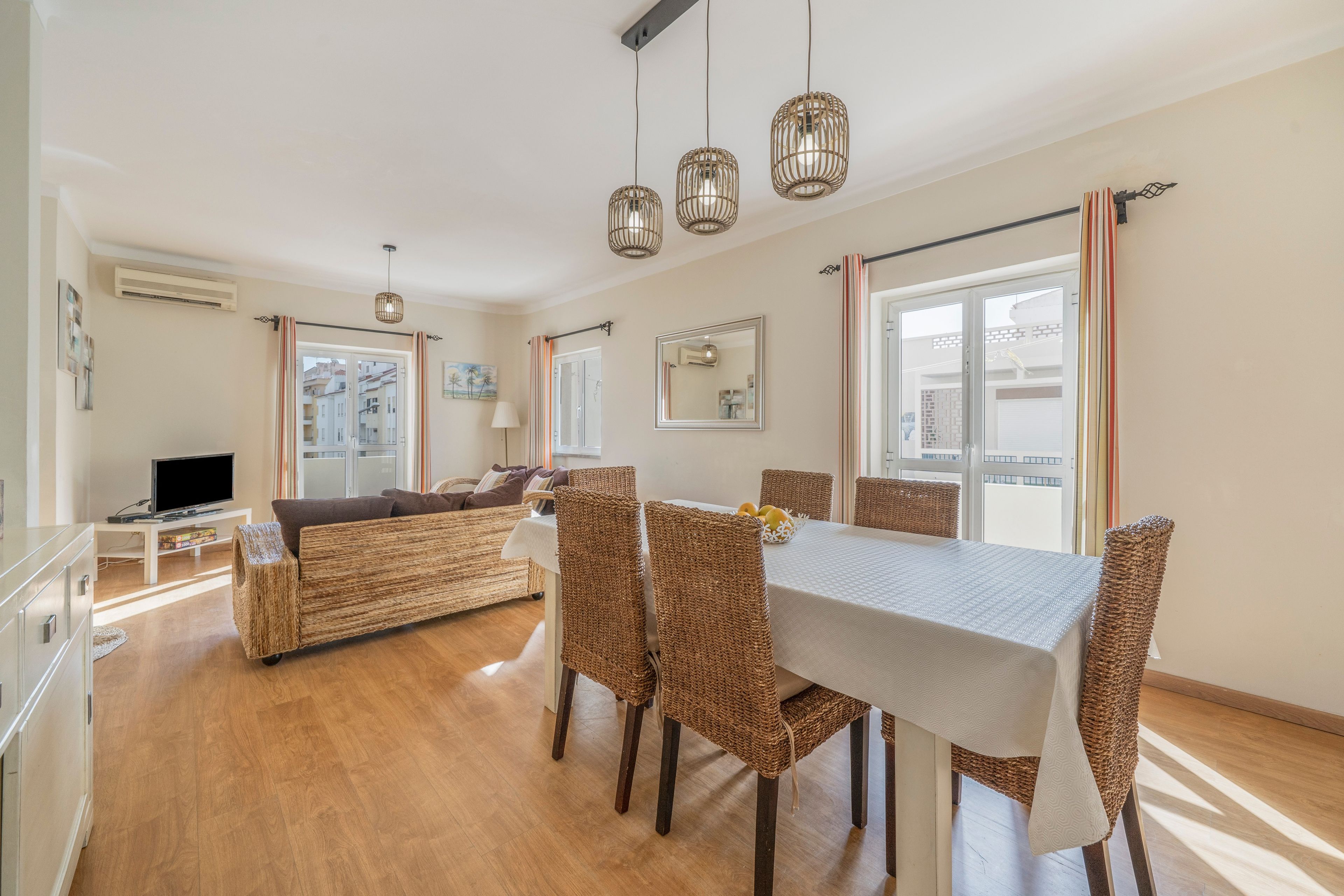 A modern living room with a sofa, dining table, wicker chairs, and a flat-screen TV.